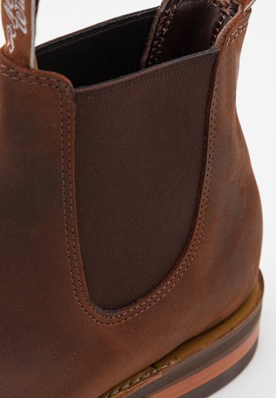 Bota Chelsea de cuero marrón con paneles elásticos laterales, suela resistente y textura suave. Presenta costuras detalladas y un acento de color contrastante.
