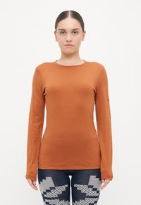 Long-sleeve, fitted top in burnt orange fabric. Round neckline, smooth texture, no patterns. Paired with blue jeans featuring a zigzag design.