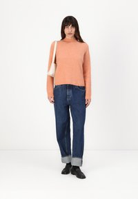 Pulover din lână de culoare portocalie deschisă, cu mâneci lungi, asortat cu blugi căptușiti albaștri, și pantofi negri. Modelul ține o geantă mică albă.