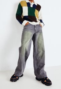 Suéter de punto con parches multicolores en verde, azul, amarillo, marrón y crema; jeans anchos grises que se desvanecen a verde en la parte inferior; zapatos negros gruesos.