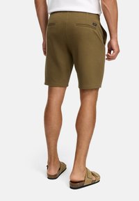 Homme portant un short vert olive jusqu'aux genoux et des sandales à enfiler beiges, debout de dos face à l'appareil photo sur fond blanc.