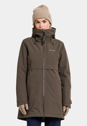 Kvinne iført en brun vinterjakke med hette, beige strikket lue og matchende hansker, stående mot en ensfarget lys bakgrunn.