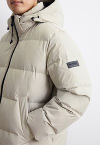 Giacca imbottita beige con cappuccio, dotata di zip frontale, polsini elasticizzati, tasche laterali e un patch con logo sul braccio sinistro. Tessuto liscio.