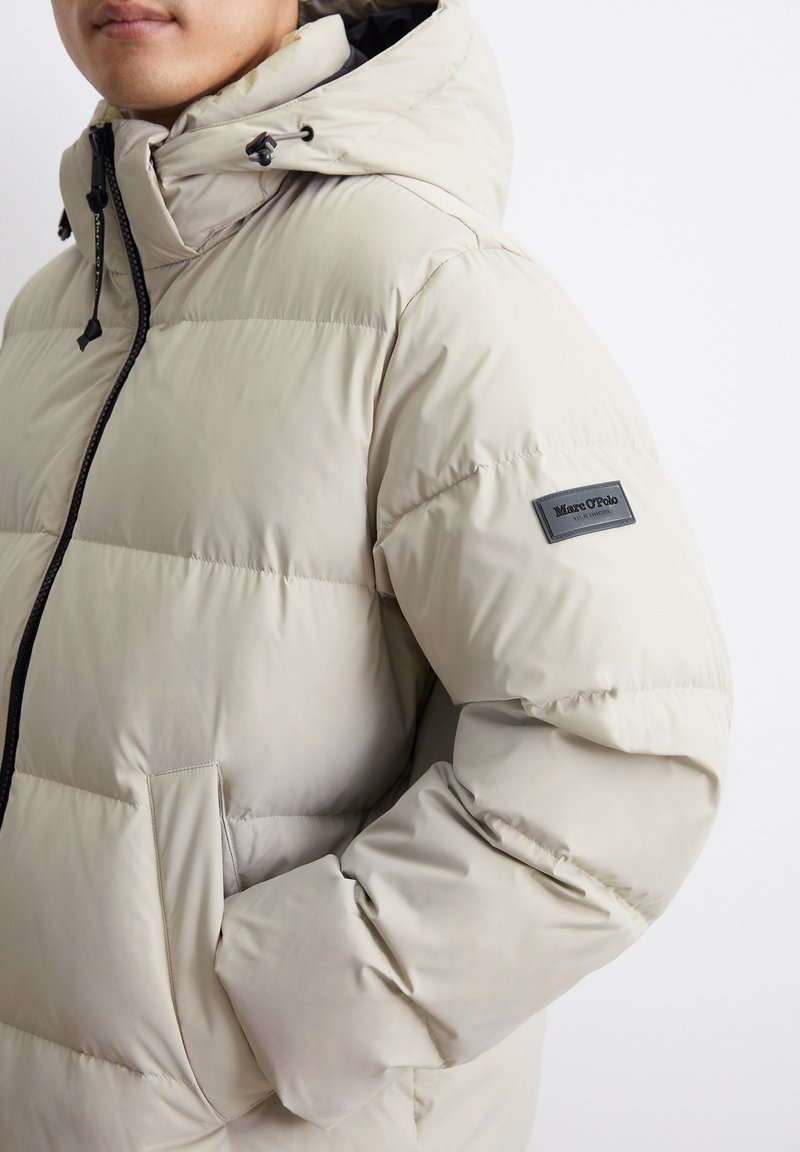 Beige gepolsterte Jacke mit Kapuze, ausgestattet mit einem Frontreißverschluss, elastischen Bündchen, Seitentaschen und einem Logo-Patch am linken Arm. Glatte Textur.