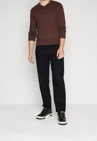 Pull marron à col en V, manches longues, associé à un jean droit noir et des baskets noires avec des accents blancs. Design simple, texture douce.