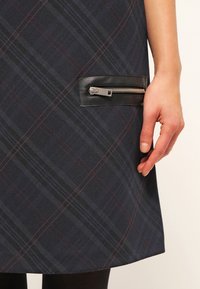 Gros plan d'une main reposant à côté d'une jupe à carreaux foncée avec une poche zippée diagonale bordée de cuir.
