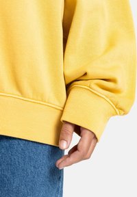 Geel sweatshirt gemaakt van zachte stof met geribbelde manchetten, met een ontspannen pasvorm. Hand rust op de zoom, in combinatie met blauwe denim.