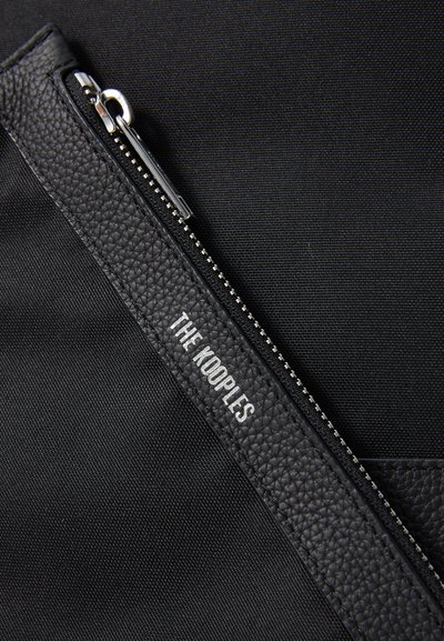 Mustapohjainen kangaspussi, jossa on teksturoitu musta nahkavetoketjun korostus ja hopeanväriset yksityiskohdat. "THE KOOPLES" on kohokuvioitu valkoisella.