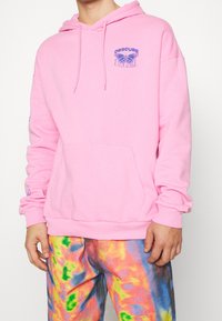 Sudadera rosa con un bolsillo tipo canguro, que presenta un gráfico de una mariposa morada y texto en el pecho. Textura suave; capucha con cordones.