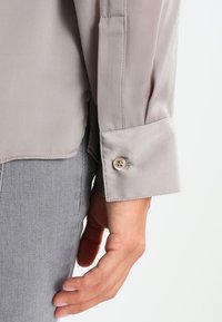 Blusa di seta grigia chiara con una texture liscia, dotata di polsino con bottoni e dettagli cuciti. Inquadratura ravvicinata dell'area del maniche e della mano.