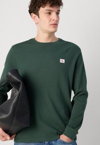 Groene, textuur rijke, lange mouwen shirt met ronde halslijn en een klein logo-patch. Model houdt een zwarte leren tas vast.