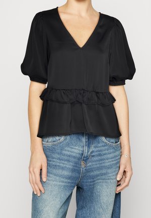 Kvinde iført en sort V-hals bluse med pufærmer og flæsedetalje ved taljen, sat sammen med falmede blå jeans.