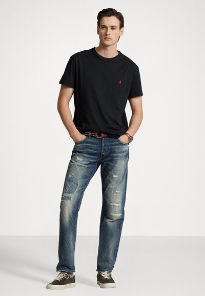 Black cotton t-shirt with a chest pocket and logo, paired with distressed blue denim jeans. Shoes are olive green with white soles.