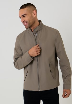 Mężczyzna uśmiechający się, ubrany w jasnobrązową kurtkę zapinaną na zamek z kołnierzem typu stojącego i zapinanymi na guziki kieszeniami z klapką, stojący na jasnym, jednolitym tle.