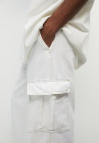 Pantalon cargo blanc en coton avec une poche latérale inclinée, des coutures visibles et une coupe décontractée. Porté avec un haut blanc ample.