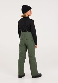 Pantaloni da neve impermeabili verdi con bretelle regolabili, due tasche posteriori e una vestibilità comoda, abbinati a una maglietta nera a maniche lunghe e un berretto.