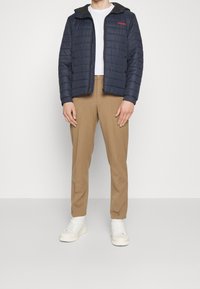 Marineblå quiltad jacka med dragkedja och huva, med en röd logotyp på bröstet, kombinerad med beige byxor och vita sneakers.