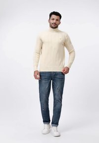 Homme portant un pull col roulé crème, un jean bleu déchiré et des baskets blanches, se tenant contre un fond gris clair.