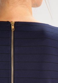 Námořnická modrá blůza s horizontálními černými pruhy a zlatým zipem na zadní straně, vyrobená z hladké, strukturované tkaniny.