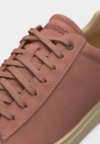 Κοντινό πλάνο σεροδοροζ σουέντ αθλητικού παπουτσιού με κορδόνια στο ίδιο χρώμα και μπεζ λαστιχένια σόλα, με χαραγμένο το όνομα της μάρκας στη γλώσσα.