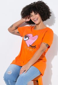 Kortærmet orange T-shirt med et pink hjerte mønster med et øje, ordet "Happy" i metallic accent. Blødt stof, afslappet pasform.