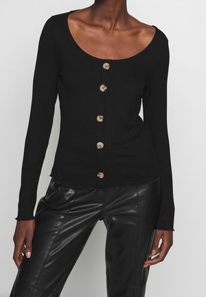Donna che indossa un top nero a coste con maniche lunghe e cinque bottoni frontali, abbinato a pantaloni in pelle nera, su uno sfondo neutro.
