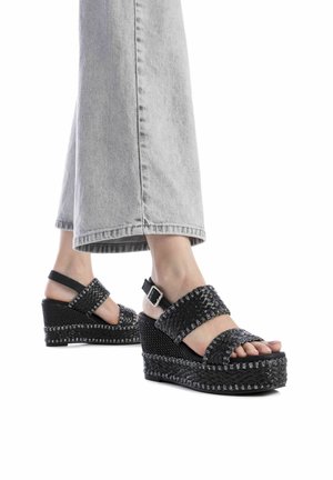 Sandali wedge neri con cinturini intrecciati, superficie testurizzata, dettagli metallici e cinturino alla caviglia regolabile. Abbinati a pantaloni palazzo grigi chiari.