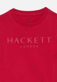 Punane puuvillane t-särk ümmarguse kaelusega. Ees on keskel heledamas toonis tekst "HACKETT LONDON".