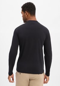 Czarna koszula z długim rękawem wykonana z gładkiego materiału. Posiada okrągły dekolt i gładkie szwy. Na zdjęciu widok z tyłu, w połączeniu z beżowymi spodniami.