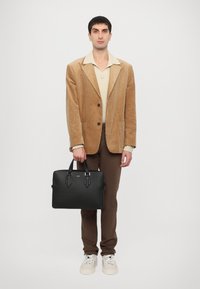 Maletín de cuero negro con doble asa, que presenta herrajes plateados y una superficie texturizada, sostenido por una persona que lleva una blazer de pana color beige.