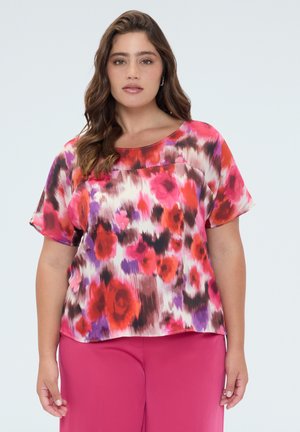Vrouw staand, gekleed in een kortarmig gebloemd bloesje met rood, roze, paars en wit patroon en felroze broek tegen een lichte achtergrond.