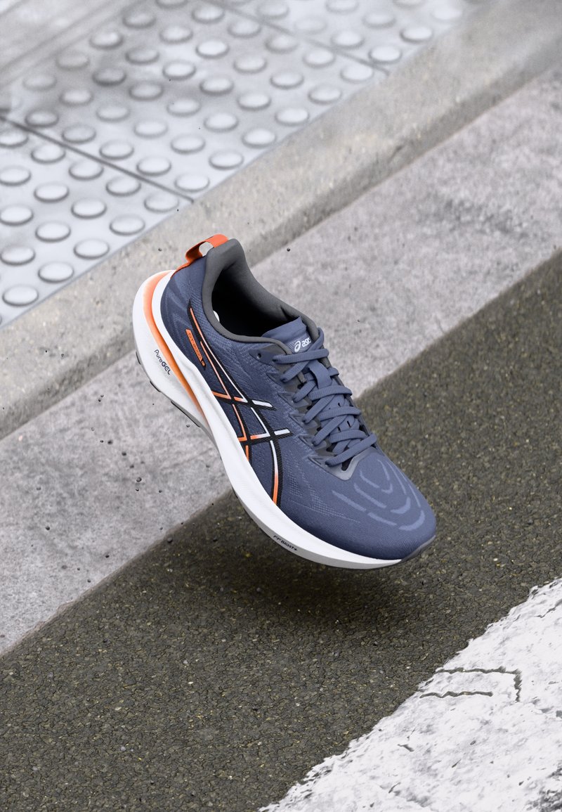 Gray athletic shoe with textured upper, orange and white accents, round toe, and cushioned sole, positioned against a textured surface.