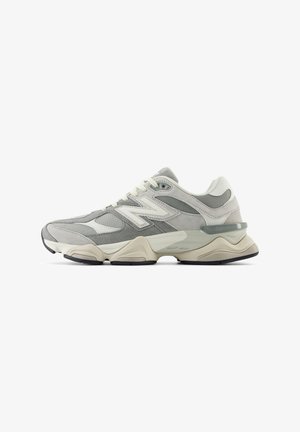 Zapatilla deportiva gris y blanca con suela gruesa, paneles de malla y ante, parte delantera con cordones y logo en el lateral, mostrada en vista de perfil.