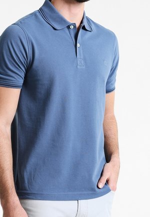 Mężczyzna w niebieskiej koszulce polo z krótkim rękawem i dwoma guzikami, w jasnych spodniach, stoi z jedną ręką w kieszeni.