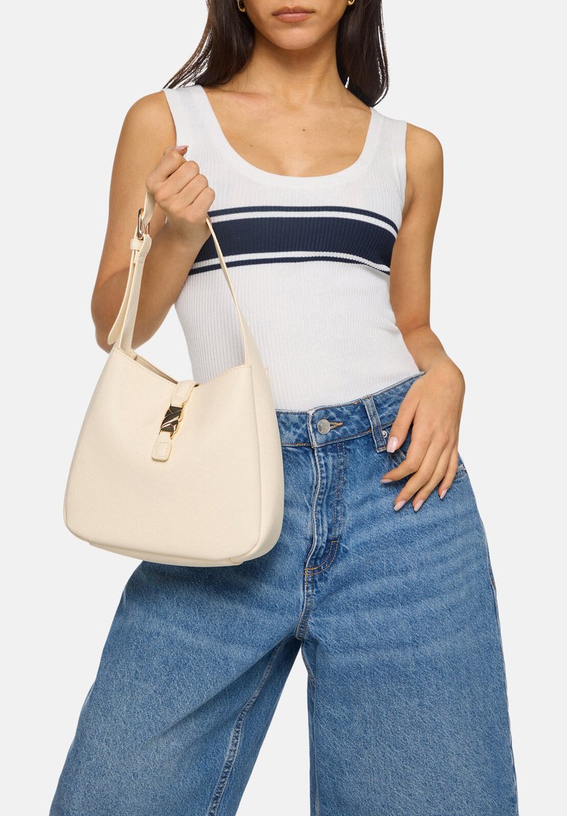 Sac à main en cuir beige avec une forme douce et arrondie, doté d'un fermoir doré et d'une seule sangle ajustable, tenu par une personne.
