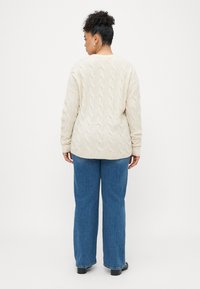 Krem pulover z cable-knit vzorcem in sproščenim krojem, s rebrastimi manšetami in robom, v kombinaciji z modrimi širokimi hlačami in črnimi čevlji.