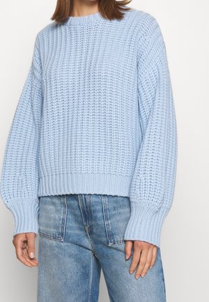 Femme portant un pull en grosse maille bleu clair avec de longues manches bouffantes, associé à un jean bleu ample.