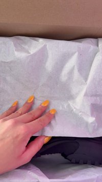 Sapatilha preta parcialmente visível sob papel de seda branco amachucado, com um cabedal texturizado e sola de borracha. Mão com unhas amarelas brilhantes segurando o papel.