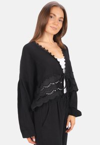 Schwarze, kurz geschnittene Strickjacke aus strukturiertem Stoff, mit gewelltem Saum und Ausschnitten sowie einer Schleifenverschlüssen an der Vorderseite. Lange Ärmel.