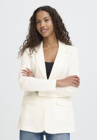 Blazer blanc au design structuré, avec deux poches avant et des revers crantés, associé à un top noir et un jean en tissu texturé.