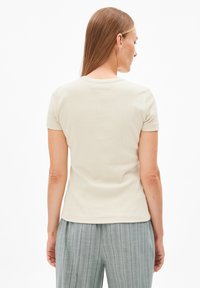 T-shirt beige chiaro a maniche corte con scollo rotondo. Tessuto a coste. Indossato con pantaloni grigi chiari a righe. Focus sulla vista posteriore.
