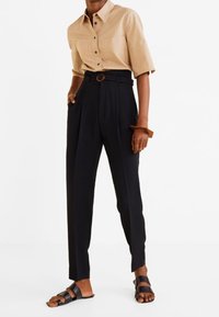 Chemise beige à manches courtes avec boutons, pantalon noir taille haute avec passants de ceinture et boucle ronde, associé à des sandales noires à brides.