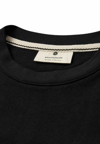 Schwarzer Sweatshirt mit Rundhalsausschnitt, der eine strukturierte Innennäht und ein kontrastierendes cremefarbenes Etikett mit schwarzem Text aufweist. Das Außenmaterial ist glatt.