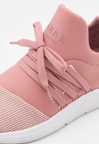 Różowe buty sportowe z teksturowanym czubkiem z siateczki i gładką tkaniną w górnej części. Posiada elastyczne sznurówki i białą gumową podeszwę. Widoczna nazwa marki.