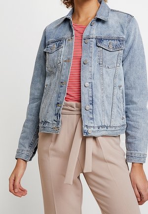 Personne portant une veste en jean bleu clair sur une chemise rayée rouge et blanche, associée à un pantalon beige taille haute avec ceinture en tissu.