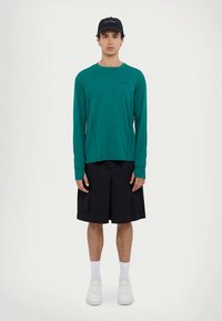 Turkio spalvos marškinėliai ilgomis rankovėmis, juodi šortai iki kelių, balti sportbačiai ir juoda kepurė. Lygi medžiaga, laisvas kirpimas ir subtilus prekės ženklo akcentas.