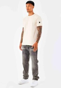 Camiseta de algodón beige clara de manga corta, con un pequeño emblema en la manga izquierda, combinada con jeans de mezclilla grises y zapatillas blancas.