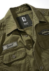 Chemise olive verte à boutons avec un écusson du logo Brandit sur la poche poitrine et un écusson Speed-and-power sur le côté droit.