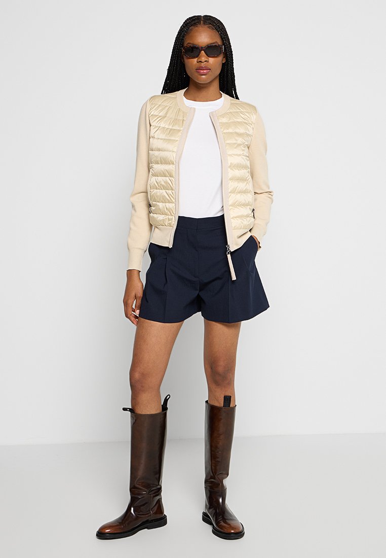Beige Daunenjacke mit gerippten Ärmeln, marineblaue Shorts und knielange braune Lederstiefel. Das Model trägt eine Sonnenbrille und steht vor einem neutralen Hintergrund.