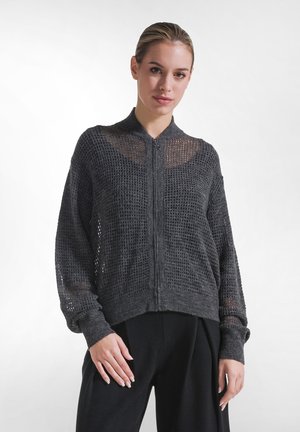 Ung kvinde med lyst hår bundet tilbage bærer en mørkegrå tekstureret sweat jakke med lynlås og løse sorte bukser, stående mod en ensfarvet baggrund.